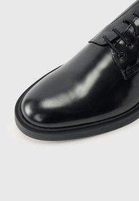 Gros plan d'une chaussure habillée en cuir noir poli avec des lacets noirs et une épaisse semelle noire, sur un fond clair.