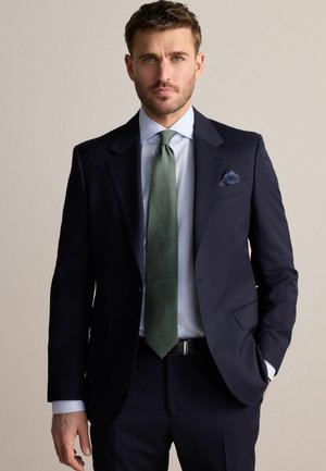 Marineblauer Nadelstreifenanzug mit aufgeschlagenem Revers, weißem Hemd, strukturiertem grünen Sale, und einem blauen Einstecktuch. Ein schwarzer Gürtel unterstreicht den Look.