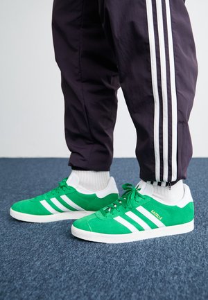 Sneaker in suede verde con strisce e dettagli bianchi, dotate di una suola testurizzata. Indossate con pantaloni neri a cono e calzini bianchi.
