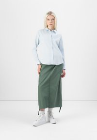 Hellblaues Hemd mit Knöpfen, grüne lange Rock mit Taschen, weiße Plateausneaker und graue Socken; minimalistisches Design, weiche Texturen.