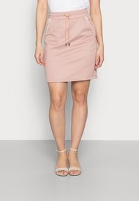 Falda rosa con un patrón, con cintura ajustable mediante cordones, que presenta dos bolsillos laterales con acentos trenzados en blanco. Combinada con sandalias de tiras de color claro.