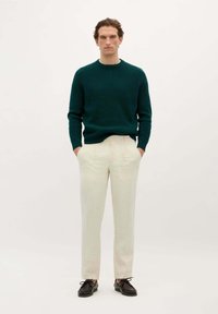 Man staat met zijn handen in de zakken, gekleed in een donkergroen sweater, crèmekleurige broek en donkerbruine schoenen tegen een effen achtergrond.