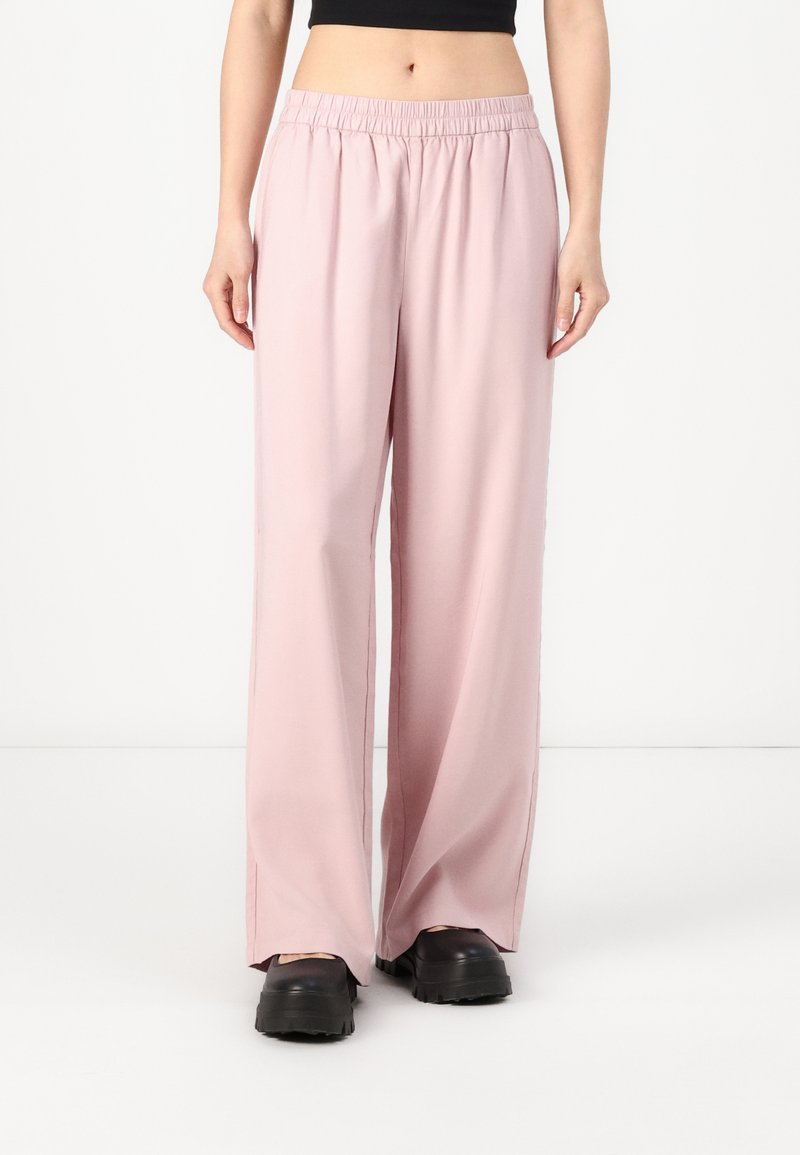 Pantaloni palazzo rosa chiaro con cintura elastica e spacchi laterali, realizzati in un tessuto morbido, abbinati a zoccoli neri.