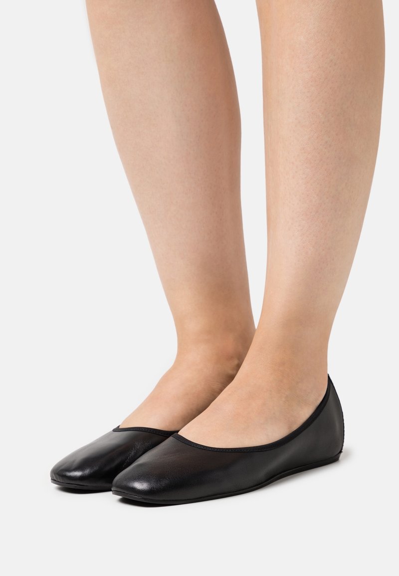 Jambes nues portant des ballerines en cuir noir simples avec des bouts arrondis sur un fond blanc uni.