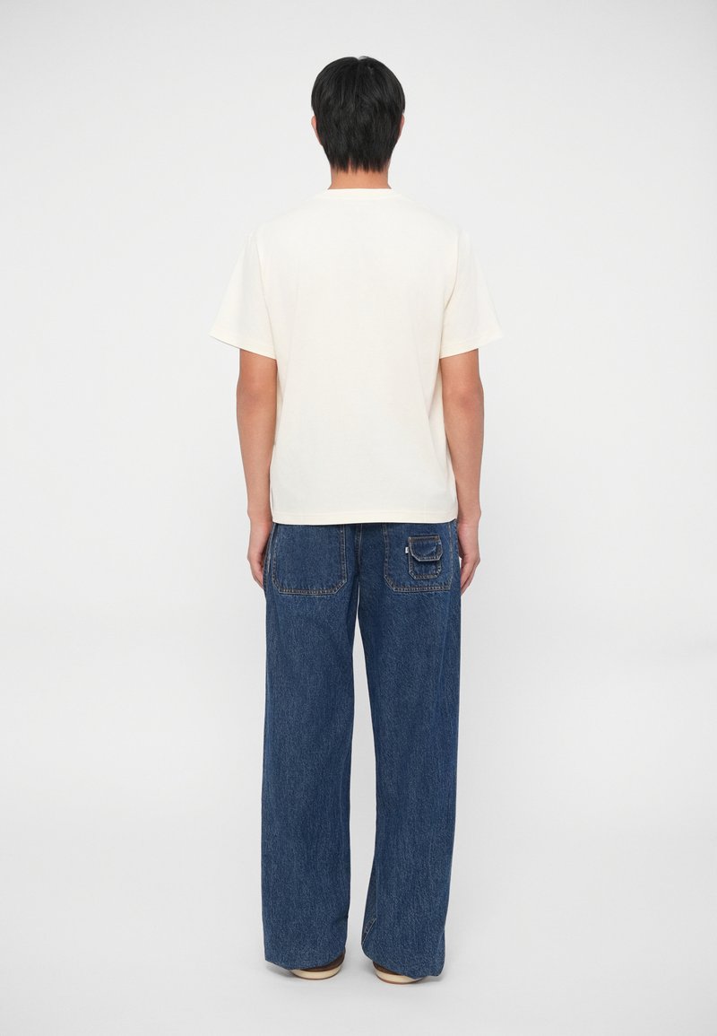 Camiseta blanca de manga corta, de corte holgado. Pantalones de mezclilla azul, de pierna ancha con detalle en el bolsillo trasero. Fondo sencillo.