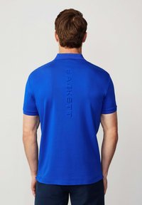 Modrá polo košile z hladkého materiálu s klasickým límcem a krátkými rukávy. Výrazné embosované logo "HACKETT" na zadní straně.