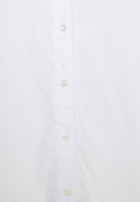 Chemise blanche à manches longues avec une texture de tissu lisse, six boutons transparents et un détail décoratif de couture le long de la patte de boutonnage.