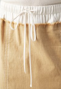 Jupe beige avec une finition texturée, dotée d'une taille élastique blanche et d'un cordon de serrage. Détail de l'ourlet effiloché qui ajoute un intérêt visuel.