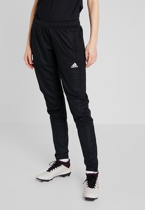 Person iført sorte Adidas træningsbukser og hvide fodboldstøvler med sorte snørebånd på en lysegrå baggrund.