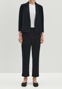 Svart skräddarsydd kavaj med två framfickor, kombinerad med åtsittande svarta byxor och en vit t-shirt med rund hals. Slät tygstruktur.