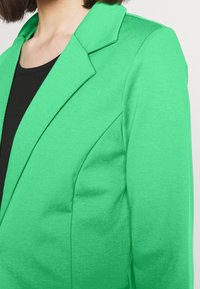 Jasnozielona marynarka wykonana z gładkiej tkaniny, z klapami z nacięciami i wyraźnie zaznaczonymi szwami na ramionach. Pod spodem widoczna czarna koszula.
