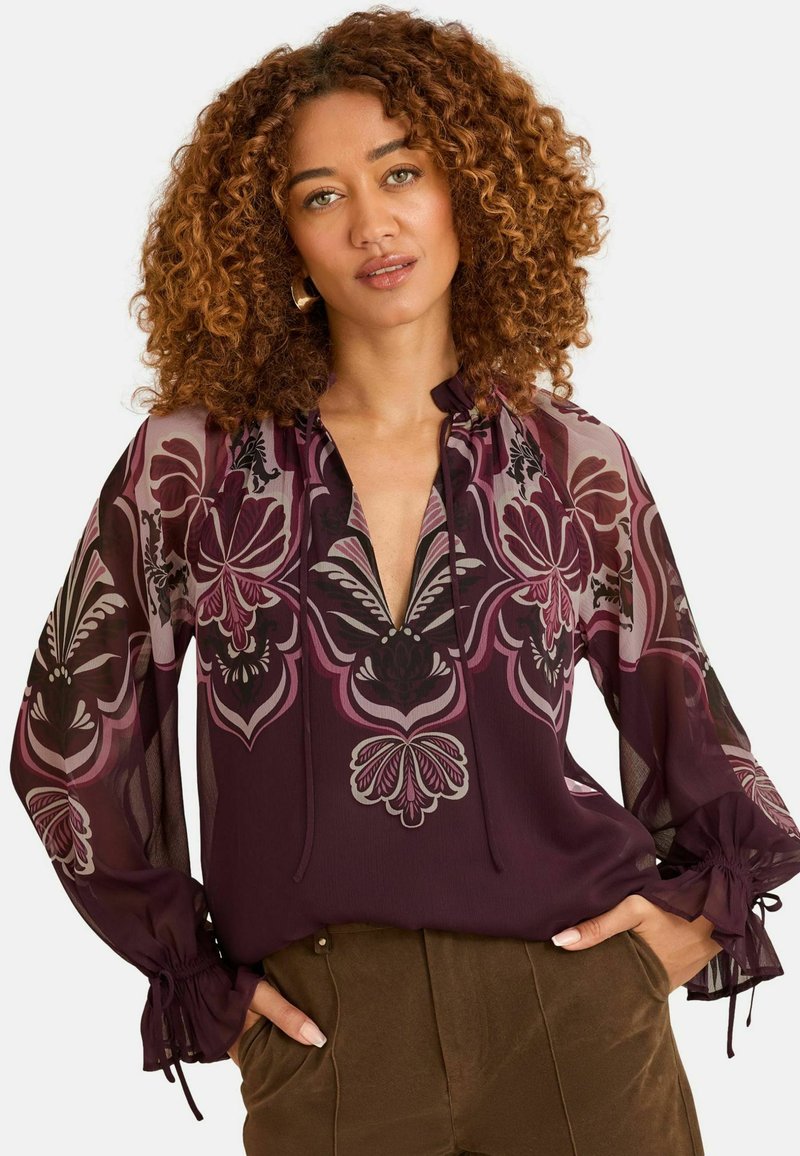 Bluse mit lila Blumenmuster, langen durchsichtigen Ärmeln, einem V-Ausschnitt und Raffung an den Bündchen. Weicher Stoff mit einem fließenden Design.
