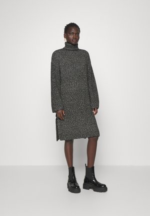 Robe à col roulé en tricot gris foncé avec des taches de couleur claire. Coupe ample, manches longues et fentes latérales. Associée à des bottines noires.