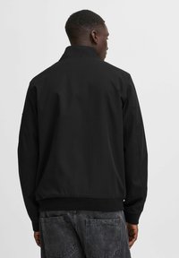Chaqueta bomber negra con cuello alzado, puños y dobladillo acanalados, que presenta una textura de tela suave y un diseño minimalista sin logotipos visibles.