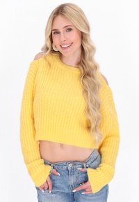 Sárga kötött, rövidített pulóver bordás textúrával és vállra csúszó dizájnnal. Hosszú ujjak széles mandzsettával, ami elnyűtt denim farmernadrággal van párosítva.