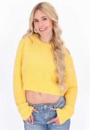 Geel gebreid cropped sweater met ribbelstructuur en off-shoulder ontwerp. Heeft lange mouwen met brede manchetten, gecombineerd met versleten denim jeans.
