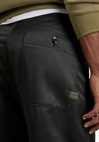 Pantalons en tissu noir avec une texture lisse. Comprend une poche arrière avec un bouton noir et une étiquette patch verte.