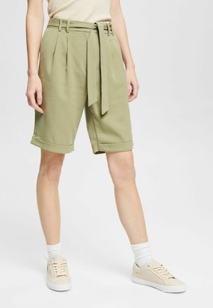 Olivengrüne, gefaltete Shorts mit passendem Gürtel, umgeschlagenen Säumen, glatter Textur, kombiniert mit beigen Sneakers und weißen Socken.