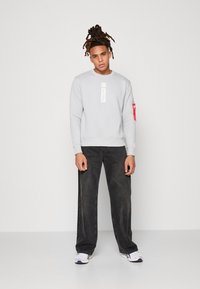 Felpa grigia con un design centrato sul logo bianco, abbinata a jeans a gamba larga grigi scuri. Presenta un accento rosso sulla manica e calzature sportive.
