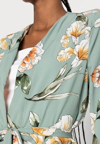 Blusa cruzada con estampado floral en tela verde con flores naranjas y blancas. Presenta un escote en V y detalle de lazo en la cintura.