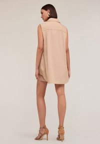 Vestito camicia senza maniche beige con colletto, vestibilità rilassata e tasche laterali. Abbinato a sandali con tacco alto a strisce di color marrone chiaro.