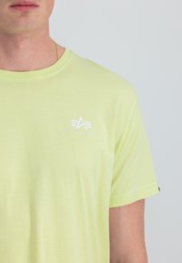 Helles gelbes kurzärmeliges T-Shirt mit Rundhalsausschnitt. Es hat ein weißes Logo auf der linken Brustseite. Weicher, glatter Stoff.