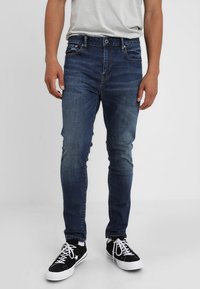 Man som bär slim-fit blå jeans, vit t-shirt, svarta strumpor och svarta låga sneakers med vita snören och sulor, stående på vit golv.