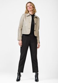 Beige leren cropped jack met een kraag en vier zwarte knopen, gedragen over een zwarte top en gecombineerd met zwarte, op maat gemaakte broek en enkellaarsjes.