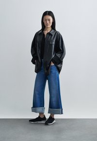 Chemise-veston oversize en cuir noir, jeans larges bleu à ourlets retroussés, et chaussures de sport noires à enfiler. Design simple et moderne.