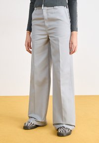 Pantalons à jambes larges en tissu gris clair avec une texture lisse, présentant une taille haute et de subtils plis avant. Chaussures à rayures noir et blanc visibles.