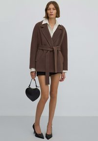 Brązowy dwurzędowy płaszcz z paskiem, o dopasowanym kroju i klapami z wycięciem. W zestawieniu z czarną torebką w kształcie serca i czarnymi szpilkami.