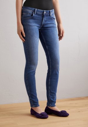 Vaqueros de denim azul ajustados con una cintura de altura media, diseño de cinco bolsillos y un desvanecimiento sutil. Se llevan con unos zapatos planos morados con un acento decorativo.