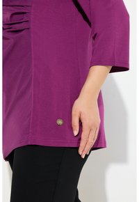 Lila Oberteil mit lässiger Passform, kurzen Ärmeln und einer gerafften Detail auf der Vorderseite. Verfügt über ein kleines Messinglogo am Saum. Schwarze Hosen sind sichtbar.