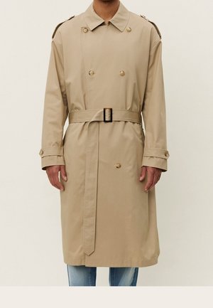 Beiger doppelt geknöpfter Trenchcoat mit Gürtel, Schulterklappen und geknöpften Manschetten, getragen über blauen Jeans.