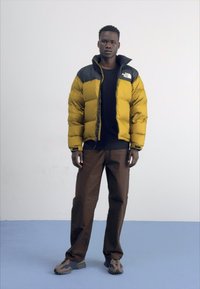 Geel en zwarte pufferjas, voorzien van een hoge kraag en ritssluiting. Bruine broek en meerkleurige schoenen maken de outfit compleet.