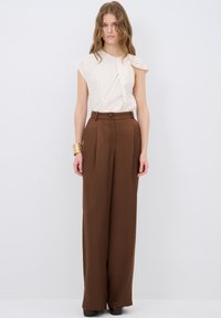 Witte blouse met korte mouwen en gedrapeerd detail, gecombineerd met high-waisted bruin wijde broek. Glad stof en een gouden armband accentueren de look.