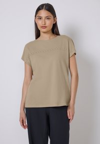 Top beige de manga corta con escote redondeado, que presenta una línea horizontal de pequeños studs decorativos en el pecho. Textura suave y corte relajado.