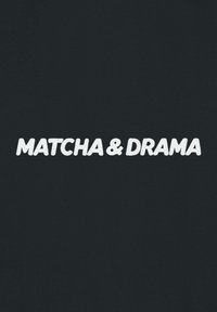 Mustaa kangasta, jossa valkoisella tekstillä lukee "MATCHA & DRAMA" lihavoidulla, pyöristetyllä fontilla, keskitettynä. Tasainen pinta ja vähäinen muotoilu.