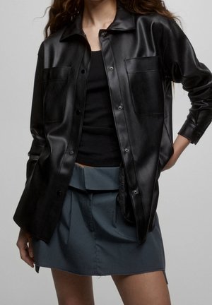 Femme portant une chemise boutonnée en similicuir noir sur un haut noir et une mini-jupe gris foncé taille haute avec détail ceinture.