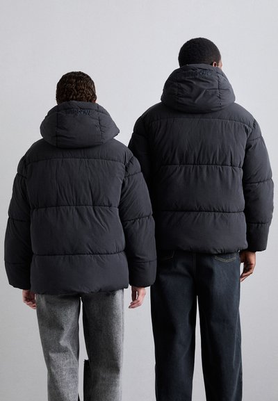 Black puffer jackets feature oversized hoods, quilted sections, and elastic cuffs, paired with gray and dark jeans. Back view.