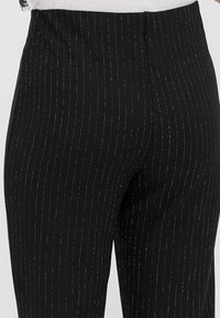 Leggings noires taille haute avec de fines rayures verticales argentées, portées avec un haut blanc rentré.
