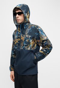 Άνδρας που φοράει σκουρόχρωμα γυαλιά ηλίου και μπλε σκούρο μπουφάν με κουκούλα με κίτρινα και μπλε σχέδια φύλλων, στέκεται με το ένα χέρι στην τσέπη.