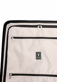 Interno del bagaglio beige con tre scomparti neri con zip e una toppa con il logo del marchio SURI FREY in bianco e nero al centro.