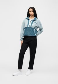 Ζακέτα από φλις σε ανοιχτό και σκούρο μπλε-πράσινο χρώμα, με φερμουάρ μπροστά, όρθιο γιακά και πλαϊνές τσέπες. Συνδυάζεται με μαύρο παντελόνι και λευκά αθλητικά παπούτσια.