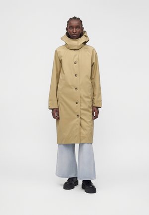 Personne portant un long imperméable beige avec capuche, un jean bleu clair à jambes larges et des chaussures noires épaisses à plateforme, debout devant un fond uni.