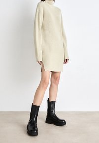 Abito in maglia a coste beige con maniche lunghe e spacchi laterali, abbinato a stivali alla caviglia neri con suola chunky, dotati di cerniera laterale e dettagli con cintura.