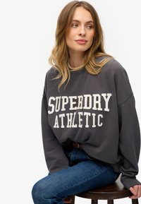 Szara bluza z napisem "SUPERDRY ATHLETIC" w białej kolorystyce, luźny krój, okrągły dekolt, ściągacze przy mankietach, w połączeniu z niebieskimi dżinsami. Gładki, casualowy materiał.