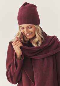 Bordeauxroter Strick-Beanie, kombiniert mit passendem Schal und Mantel, zeigt weiche Texturen und ein lässiges, lässig drapiertes Design.