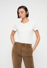 Άσπρο μπλουζάκι με κοντά μανίκια και στρογγυλή λαιμόκοψη, σε συνδυασμό με καστανά παντελόνια από κοτλέ ύφασμα. Το ύφασμα φαίνεται μαλακό με διακριτική υφή.