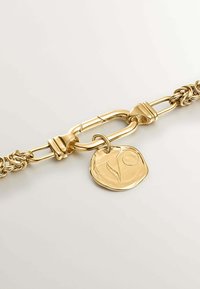 Pulsera de cadena de oro con un gran eslabón central y un colgante redondo con una superficie texturizada y diseño grabado. Acabado suave en toda la pulsera.